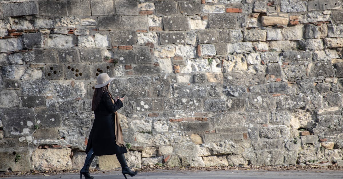 Stylish woman in trench coat and hat walking past an ancient stone wall.
