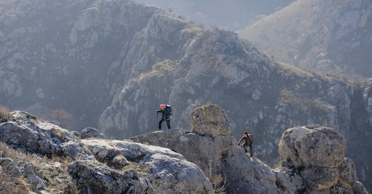 Two hikers traverse a rugged rocky terrain in a mountainous landscape under clear skies.