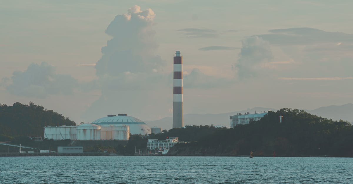 Industrial skyline featuring a smokestack by a serene lake under a clouded sky.