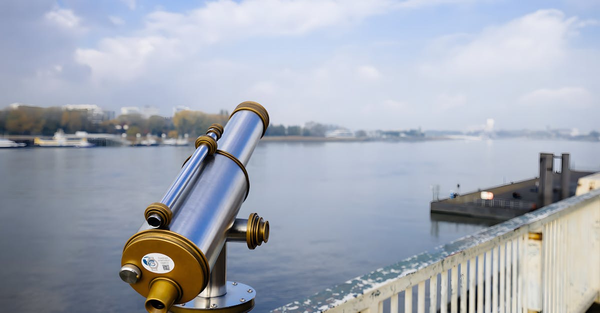 Fixed telescope on a railing, facing a serene urban waterfront under a cloudy sky.