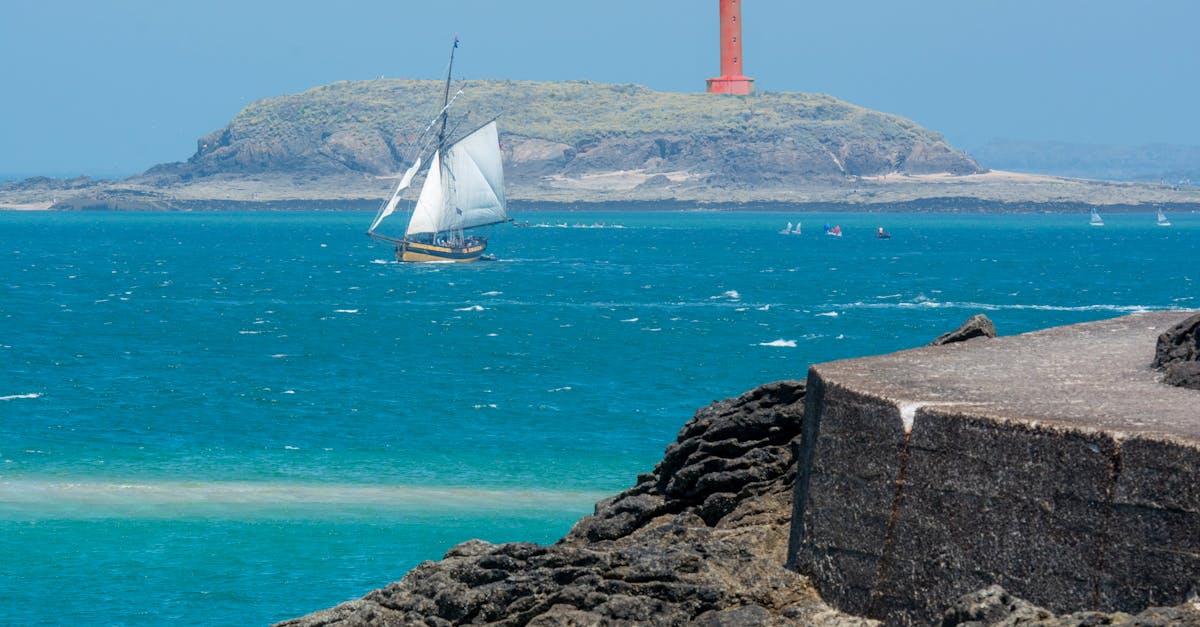 Sailboat gliding on turquoise waters near a red lighthouse on a rocky island coast.
