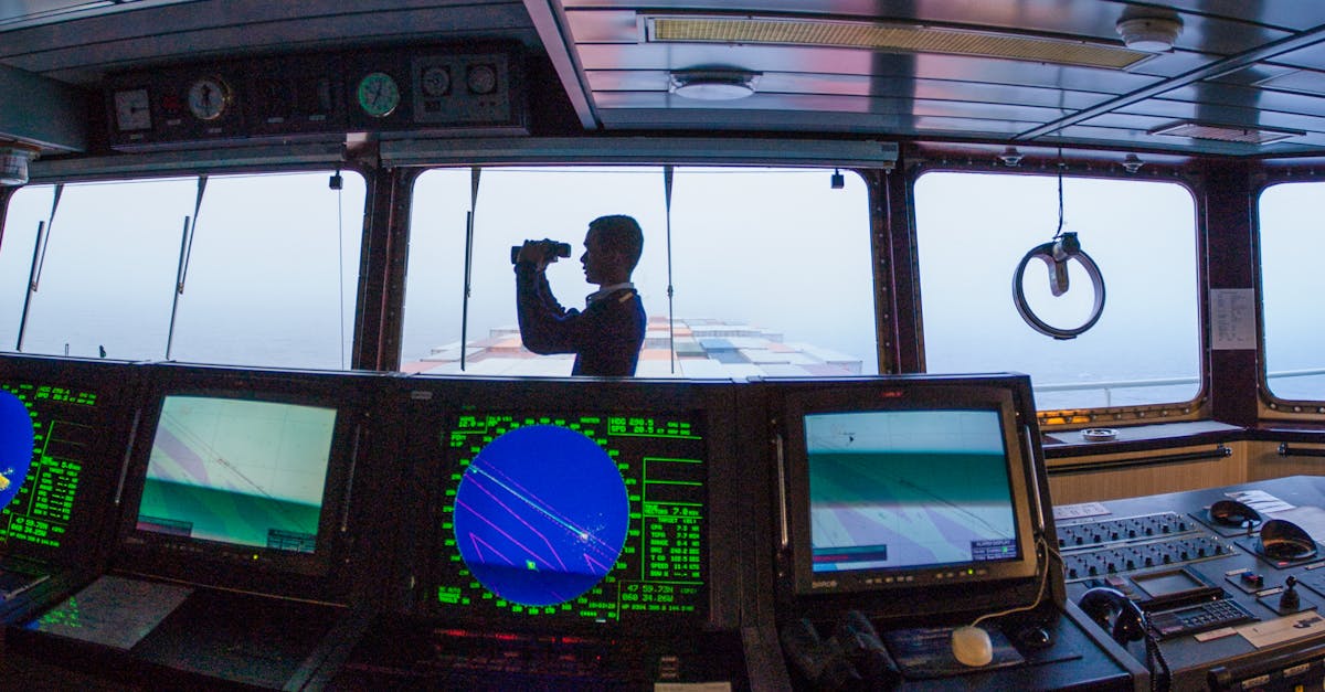 Silhouette of a navigator using binoculars on a cargo ship's bridge, surrounded by navigation equipment.