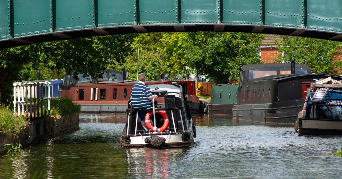 A canal boat navigates under a green bridge on a sunny day with lush surroundings and other boats docked nearby.