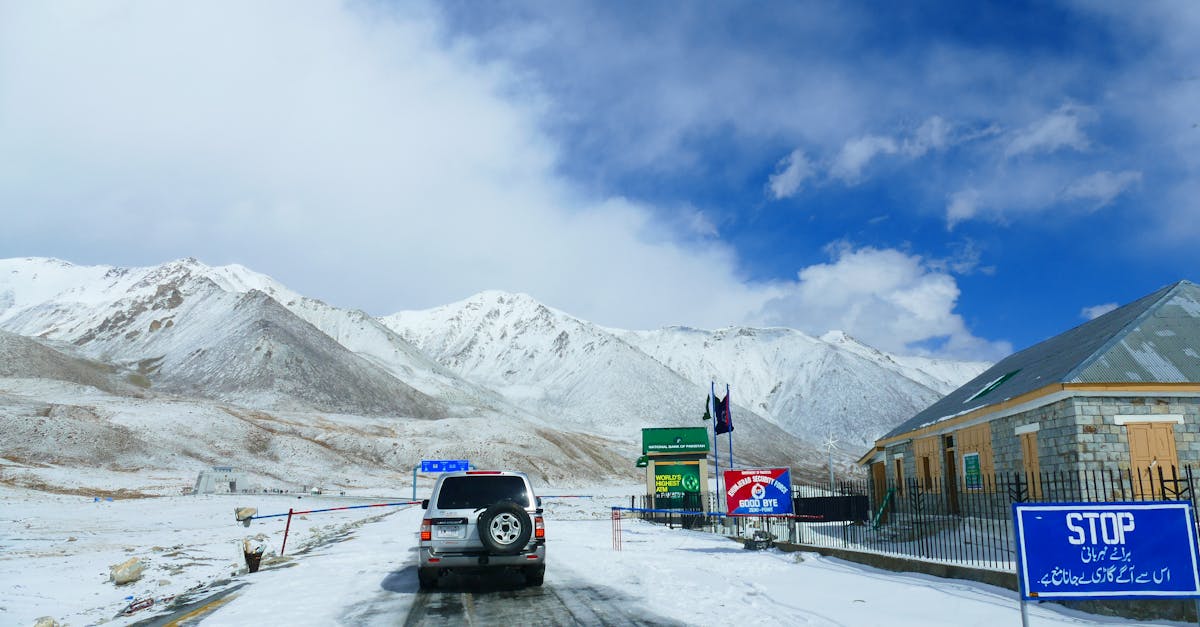A vehicle approaches a snow-covered mountain pass with border signs and snowy peaks in the background.
