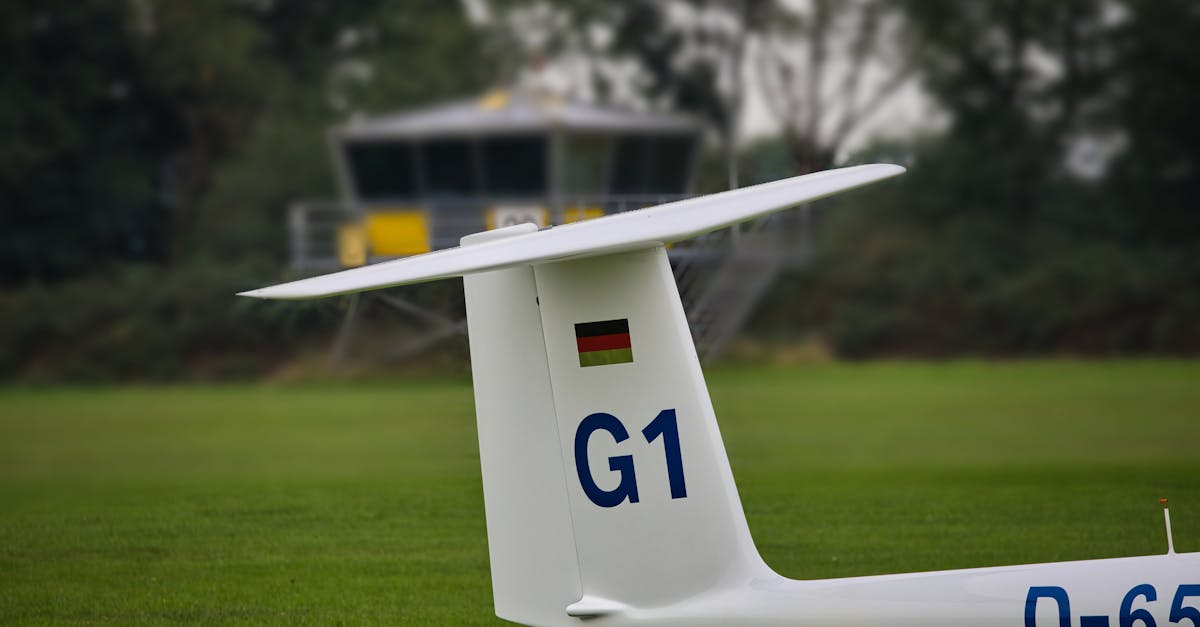 Close-up of a glider's tail with a control tower in the background at a rural airfield.