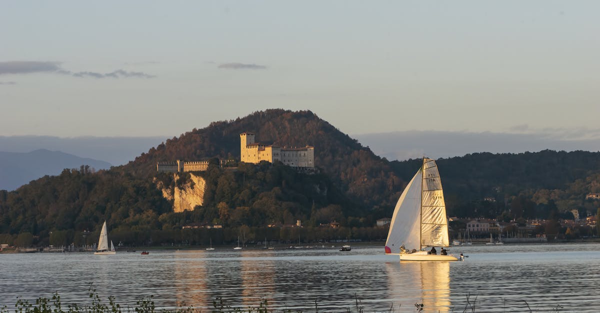 A serene sailboat glides on Lake Maggiore at sunrise, with Rocca Borromeo in the background.