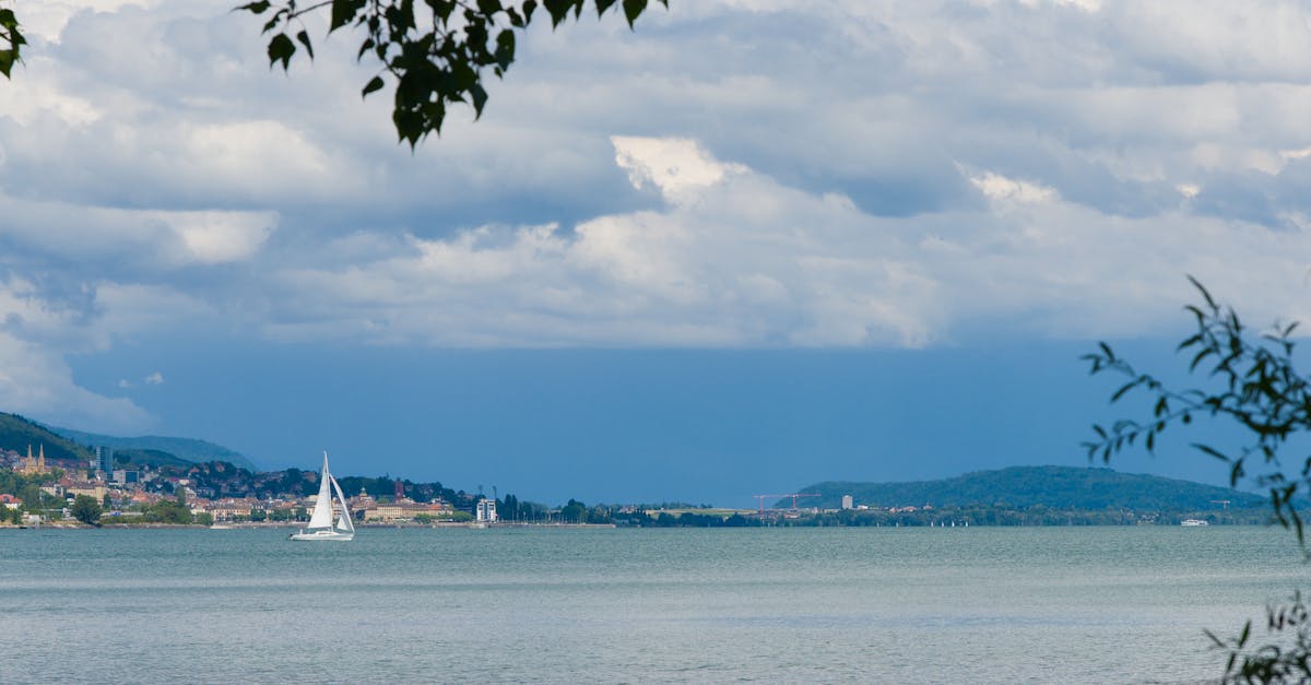 Serene lake view with sailboat navigating under a dramatic cloudy sky and distant cityscape.