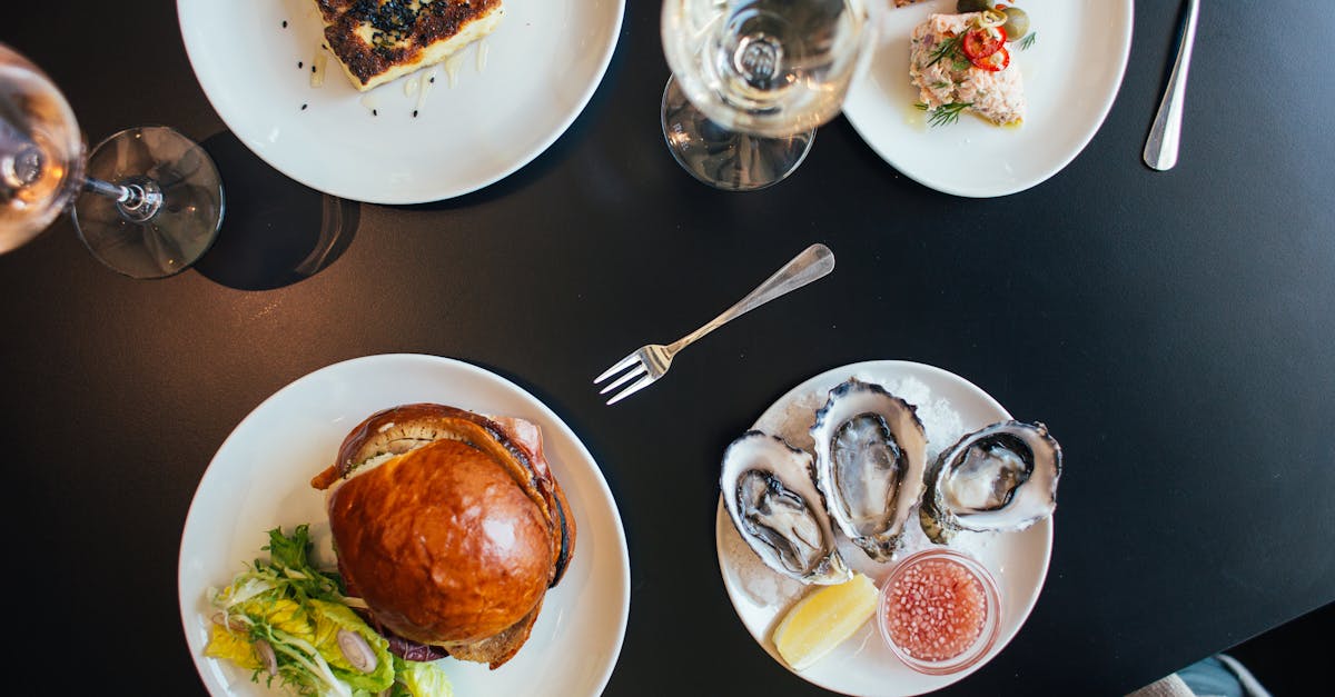 Top view of black table with plates with burger with herbs near pie with poppy seeds near crackers and mussels near bowl with sauce and forks near glasses with wine