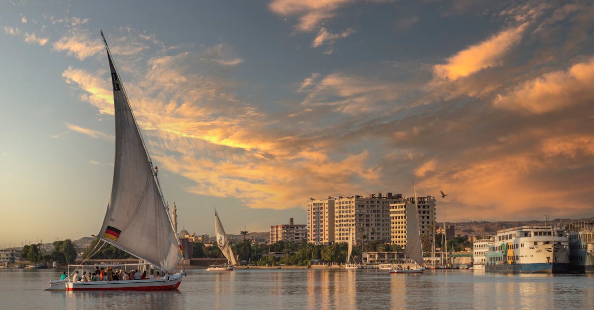 A traditional felucca sails the Nile at sunset with cityscape in the background.