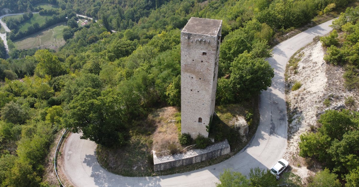 Drone view of ancient tall stone tower surrounded by rural road loop and located on verdant forested hill slope on sunny summer day