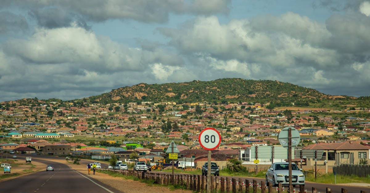 A vibrant countryside town nestled by mountains and a scenic road with vehicles under a blue sky.