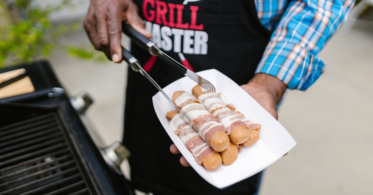 A person in an apron grilling bacon-wrapped hotdogs outdoors on a sunny day.