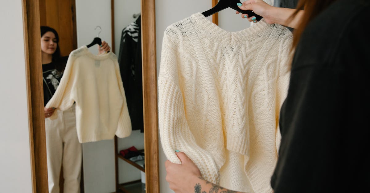 Woman holding and trying on a knitted sweater while observing her reflection in a mirror, showcasing fashion styling.